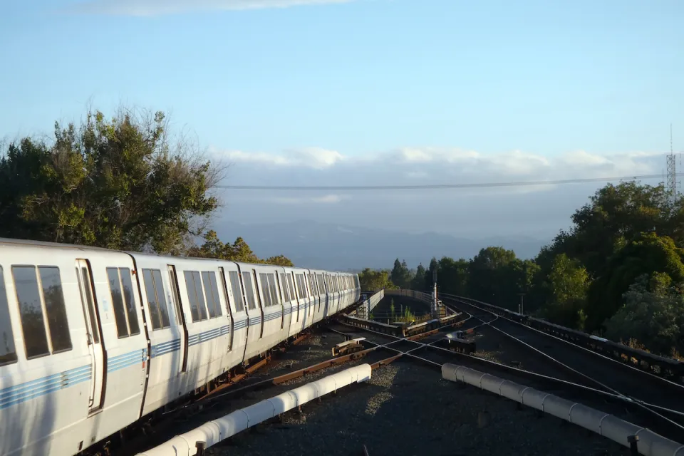 BART heavy rail train on elevated track pulling into Concord, CA station with cloudy sky and trees in background.